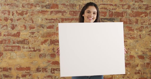Portrait of happy biracial woman with long hair holding white board with copy space by brick wall. Lifestyle, mock up, message and information, unaltered.