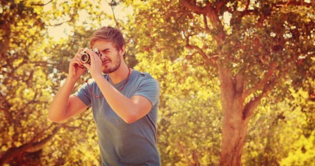 A young Caucasian man is capturing a moment with a vintage camera outdoors, with copy space. His focused expression and casual attire suggest a relaxed photography session in a natural setting.