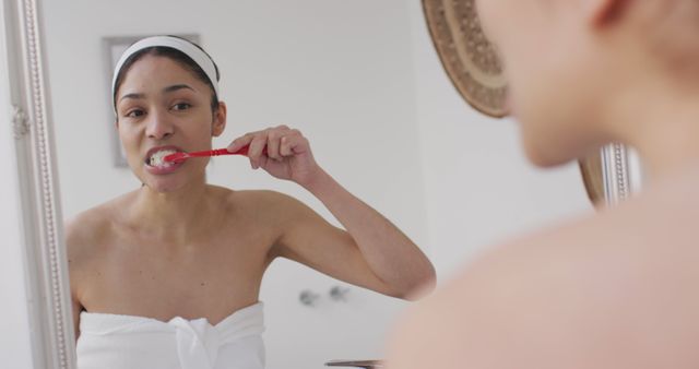 Woman in bathroom brushing teeth while looking in mirror wrapped in towel. Perfect for promoting dental care products, personal hygiene routines, health and wellness campaigns, and skincare advertisements.
