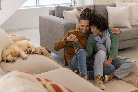 Couple relaxing on living room floor sharing smartphone while puppy sleeping on sofa