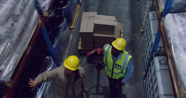 High angle view of two warehouse workers wearing yellow hard hats inspecting inventory on a loft aisle. One worker is pointing while the other observes several boxed packages and storage items placed on shelves. Ideal for use in articles, blogs, and manuals about warehouse management, logistics, supply chain, team collaboration, and industrial safety.