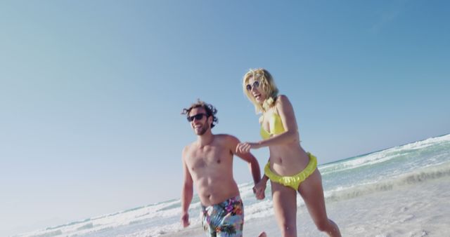 Couple in swimwear enjoying a sunny day at the beach, holding hands while running along the shoreline. Ideal for promoting travel and tourism, summer vacations, leisure activities, and romantic getaways. Represents happiness, togetherness, and relaxation.