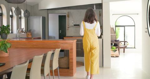 Mature woman walking toward modern wood kitchen island in yellow overalls, minimalist daylight inter