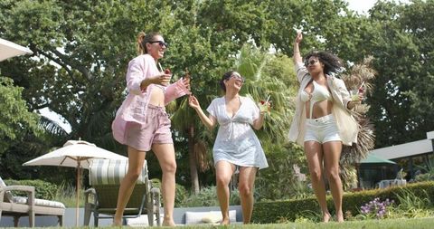 Diverse women dancing on resort lawn holding cocktails in swimwear and sunglasses
