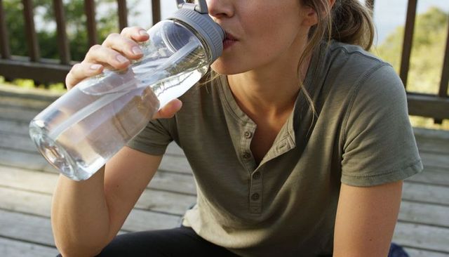 Woman drinking from reusable water bottle on wooden deck porch enjoying outdoor hydration