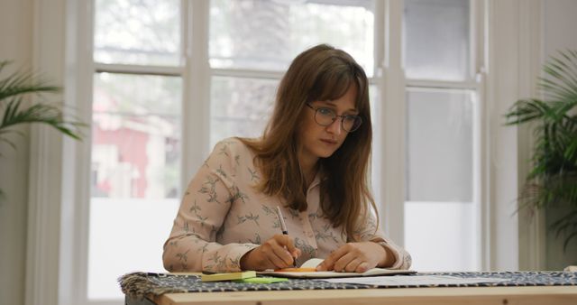 Focused Woman Writing on Notebook at Desk with Large Windows - Download Free Stock Images Pikwizard.com