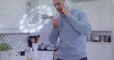 Man tasting food while talking on smartphone with ar interface overlay in modern kitchen