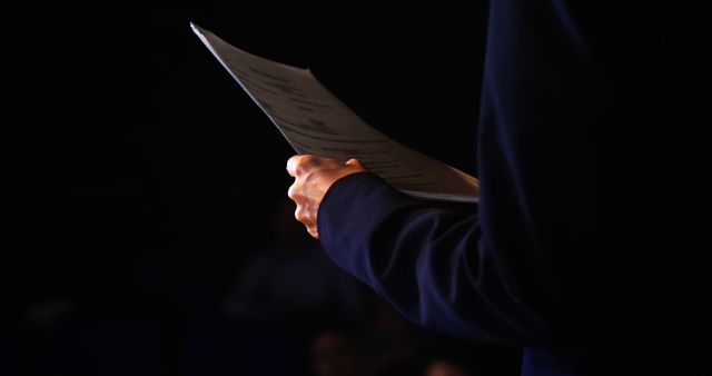 Person is holding papers which suggest they are prepared to deliver a speech or presentation on stage. Dark background adds focus on the presenter. Perfect for themes like public speaking, business presentations, seminar preparations, or lectures. Ideal for use in articles discussing presentation skills, effective communication, business training, and educational seminars.