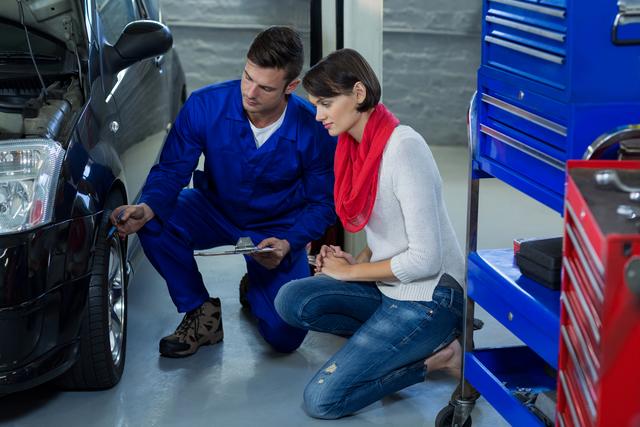 Mechanic in blue coveralls showing a female customer an issue with her car in an auto repair shop. Ideal for use in articles about car maintenance, automotive repair services, customer service in the automotive industry, and professional mechanical diagnostics.