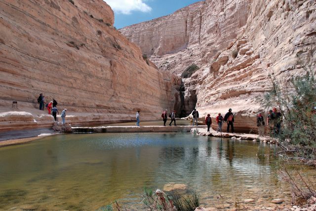 Tourists are seen exploring and hiking through a magnificent desert canyon in Israel. The canyon features dramatic rock formations and a serene pool of clear water, offering stunning natural beauty. This image can be used for travel blogs, tourism advertisements, nature exploration promotions, and adventure print materials showcasing the breathtaking landscapes and outdoor activities available in Israel.