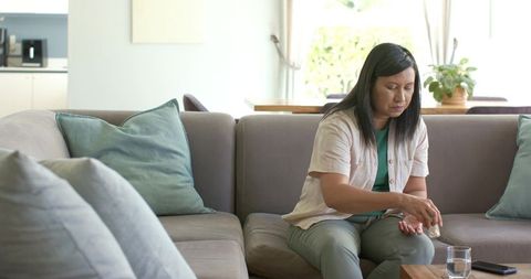 Asian woman taking pill while sitting on sectional sofa at home with glass of water