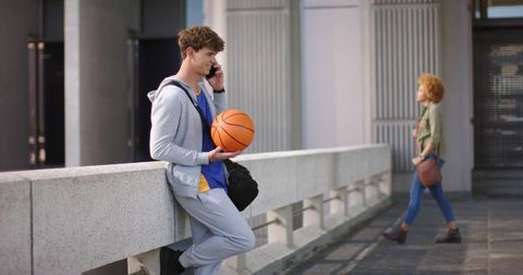 Young man leaning on railing holding basketball talking on phone urban campus lifestyle