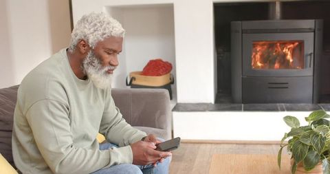 Senior man relaxing on sofa using smartphone by fireplace
