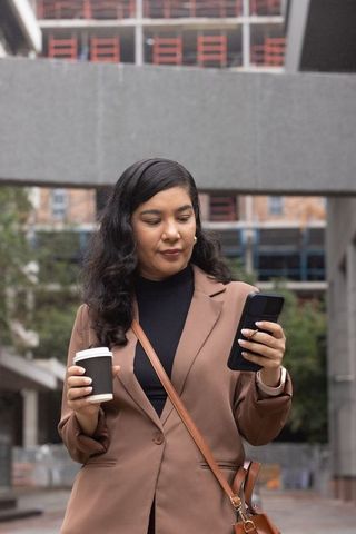 Professional woman checking smartphone and holding coffee in urban plaza with scaffolding