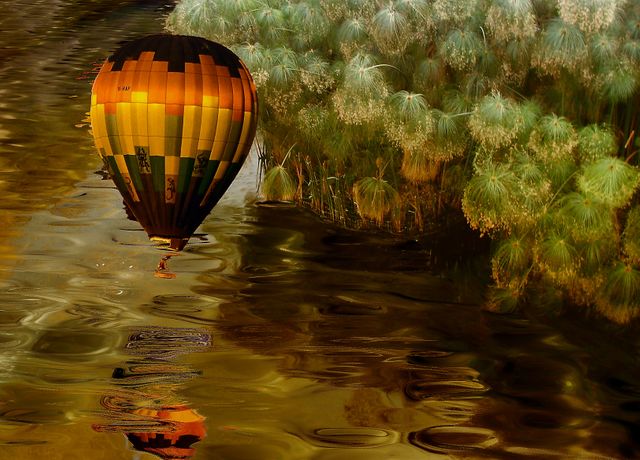 Hot air balloon floating over calm river with a beautiful reflection, surrounded by lush greenery and illuminated by warm sunset light. Perfect for projects involving travel, adventure, nature exploration, calm and relaxation themes.