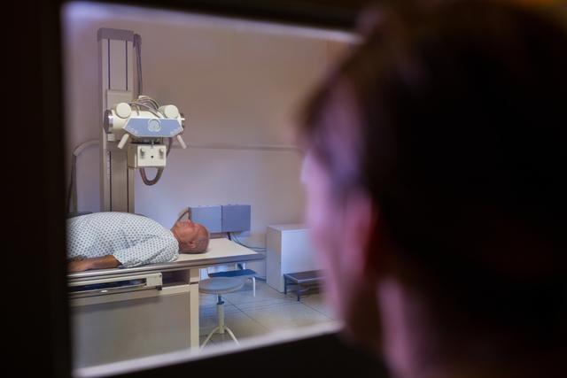 This image depicts a male patient lying on an examination table while undergoing an x-ray test in a hospital. The scene includes medical equipment and a healthcare professional observing through a window. This image can be used in articles or materials related to healthcare, medical diagnostics, patient care, and hospital procedures.