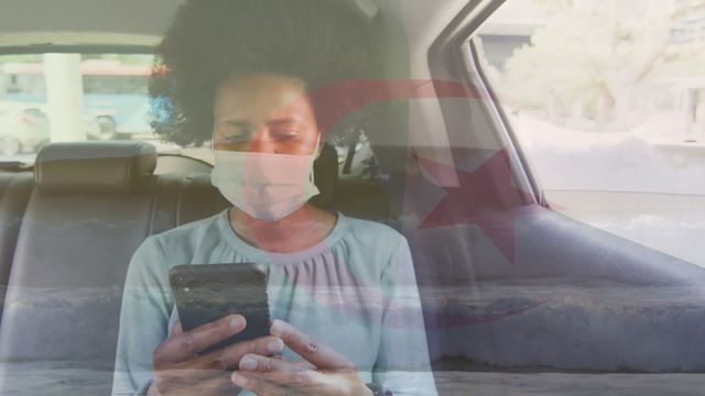 An African American woman wearing a face mask uses her smartphone while sitting in a cab, with the Algeria flag digitally overlaid in the video. Ideal for concepts related to health and safety during the COVID-19 pandemic, use of public transportation, technology in daily life, and patriotism. Useful for articles, blog posts, and social media content centered around public health measures, traveling safely, or national pride during the pandemic.