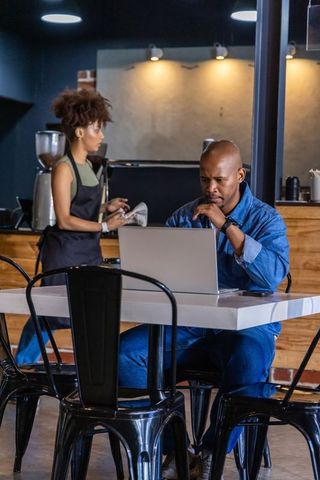 African american man working on laptop in modern cafe while barista drying cups