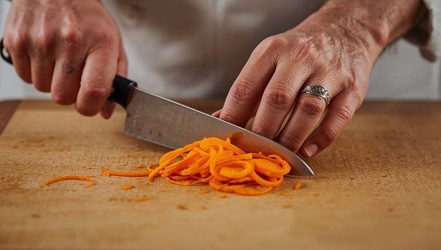 Chef slicing carrot ribbons with chef knife on wooden board closeup, culinary prep