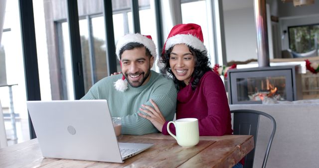 Couple wearing Santa hats enjoying a Christmas celebration through video call on a laptop. They are smiling and sharing a happy moment together. A cozy setting with a fireplace in the background adds festive warmth. Ideal for themes of remote holiday celebrations, virtual gatherings, and togetherness during the Christmas season.
