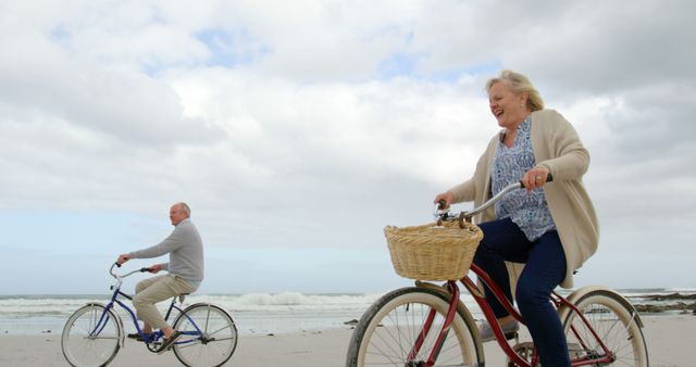 A senior couple riding bicycles along a sandy beach with the ocean waves and cloudy sky in the background. They appear to be having fun and enjoying their leisure time while maintaining a healthy lifestyle. This image can be used for promoting active retirement, healthy lifestyles for seniors, outdoor activities, and travel destinations.