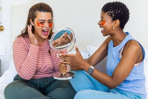 Two friends laughing during skincare night wearing under-eye gel patches holding mirror