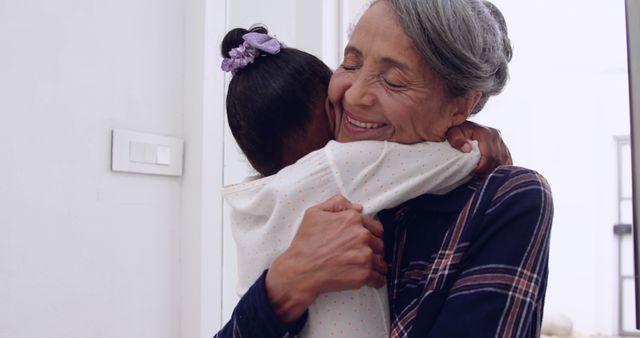 This image shows a joyful grandmother embracing her granddaughter in a loving hug inside a bright home. The joyful expression of the elderly woman and the affectionate embrace convey strong family bonds and emotional connection. This image can be used in advertisements, social media posts, or articles that focus on family, intergenerational relationships, caregiving, or home life.