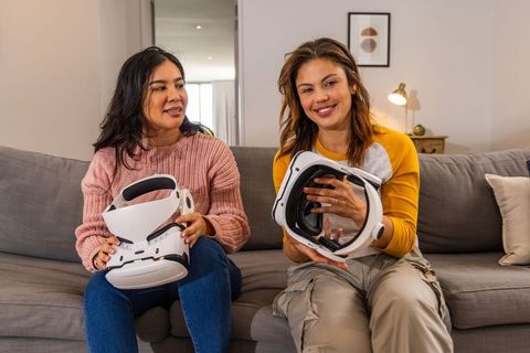 Female friends sitting on sofa holding vr headsets enjoying virtual reality at home