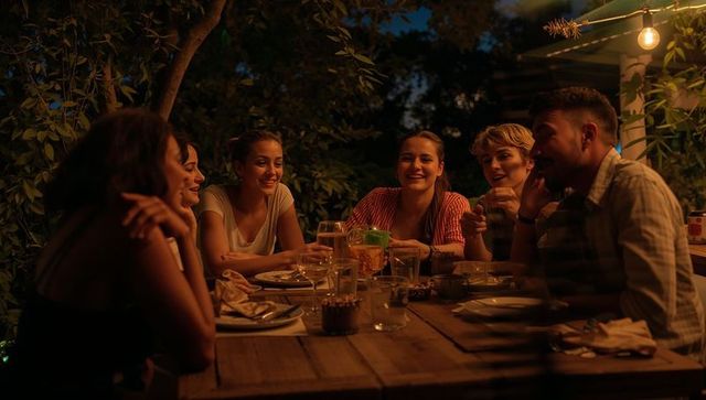 Laughing friends around rustic wooden table under string lights sharing drinks outdoors