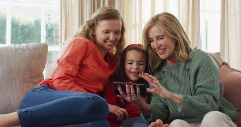 Multigenerational women sharing smartphone on cozy sofa, laughing in bright living room