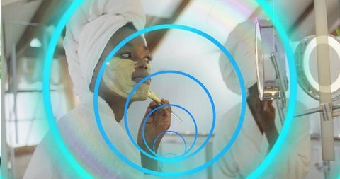 Applying facial mask at vanity wearing towel turban and bathrobe with neon mirror rings