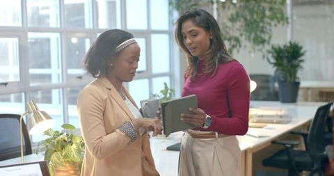 Two professional women collaborating on tablet in bright modern office with coffee mug