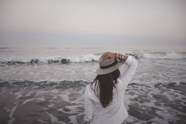 This stock photo depicts a woman standing on a beach and looking at the ocean waves during sunset. She is wearing a white shirt and hat, her back turned to the camera, suggesting a moment of relaxation and reflection by the sea. Ideal for use in travel blogs, wellness articles, or lifestyle websites to convey themes of tranquility, escape, and nature appreciation.