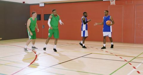 Four men playing basketball indoors, green team driving past blue defenders during pickup game