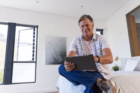 Middle-aged man relaxing on bed using tablet in modern bright minimalist bedroom