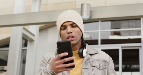 Young man checking smartphone at glass building entrance wearing beanie and headphones