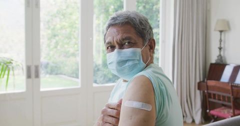 Senior man wearing surgical mask showing vaccination bandage at home after shot