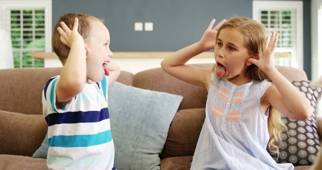 Two kids are making funny faces while sitting on a couch at home. They seem to be enjoying themselves and sharing a playful moment. This can be used to depict family bonding, sibling relationships, or a fun home environment. Perfect for family-oriented advertising, children's products, or articles about family dynamics or playtime.