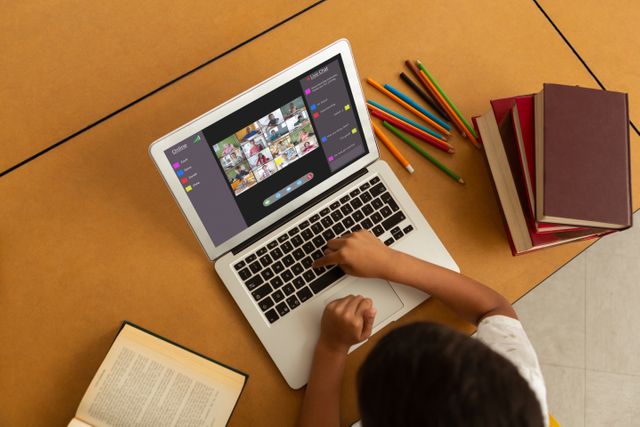 Child using laptop during online school lesson, interacting with other students remotely. Surrounding items like notebooks, books, and colored pencils suggest an engaging and creative learning environment. Ideal for illustrating online education, remote learning tools, homeschooling, or technology in education.