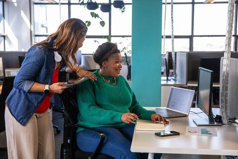 Diverse women collaborating in modern open-plan office mentoring coworker using laptop