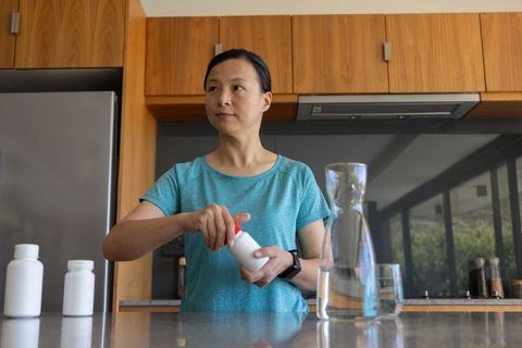 Asian woman opening supplement bottle at kitchen counter with water carafe