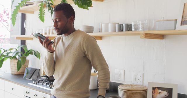 Man standing in a modern kitchen, speaking into a voice assistant on his smartphone. Shelves with plants and kitchen items on the wall. Ideal for illustrating smart home technology, modern lifestyle, or technology use in the household contexts.