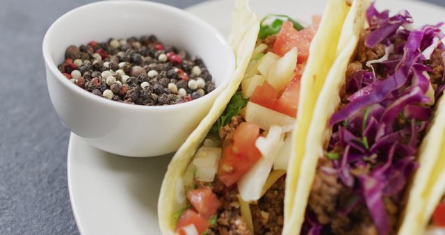 Freshly made tacos with various fillings including ground meat, lettuce, tomatoes, onions, and red cabbage. Nearby bowl of mixed peppercorn seasoning. Ideal for use in restaurant promotions, culinary blogs, recipe websites, and food product advertisements showcasing authentic Mexican cuisine.