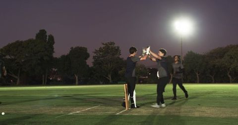 Floodlit cricketers celebrating after wicket on grass pitch at dusk showing teamwork
