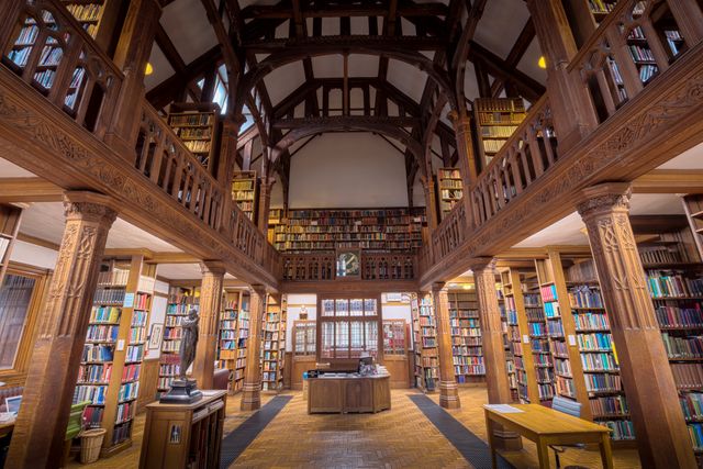 This image features a grand library with a wooden interior, showcasing an extensive collection of books arranged on shelves reaching up to a high ceiling, supported by wooden beams. Ideal for use in educational or historical publications, illustrated articles on reading culture, academic brochures, websites promoting libraries or knowledge institutions, and visual content emphasizing traditional and vintage aesthetics.