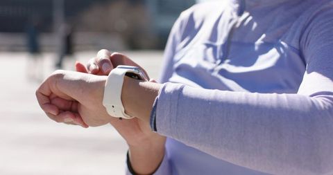 Woman adjusting white-banded smartwatch on wrist while standing outdoors in purple athletic top