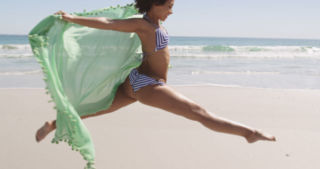 Young woman, wearing striped bikini and green cover-up, leaping gracefully on sunny beach. Waves in the background, clear sky. Perfect for promoting summer vacations, beachwear, body positivity, active lifestyle.