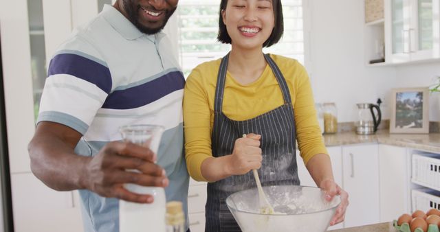 Interracial couple having fun while baking together in kitchen, mixing ingredients in bowl and smiling. They wear casual clothes and an apron, enjoying domestic time bonding in home kitchen. Perfect for themes like relationship bonding, culinary joy, home life, and multicultural lifestyles.