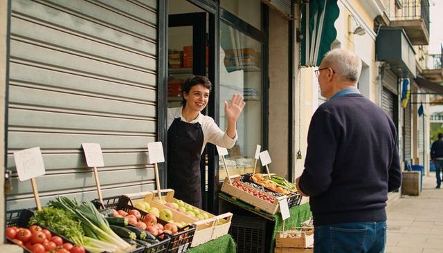 Friendly market vendor waving to customer at shop doorway with fresh produce crates