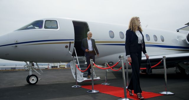 Businessman with grey hair and businesswoman with curly blond hair disembarking a private jet. Red carpet setup, signifying luxury travel. Ideal for illustrating executive travel, business success, professional lifestyle, corporate events, and luxury services.